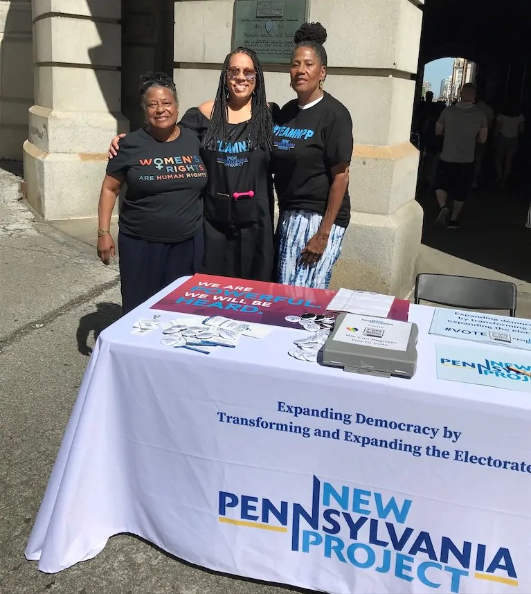 Jovida (izquierda) con miembros de la comunidad afuera del Ayuntamiento en Filadelfia. Están parados detrás de un cartel del Proyecto Nueva Pensilvania. 