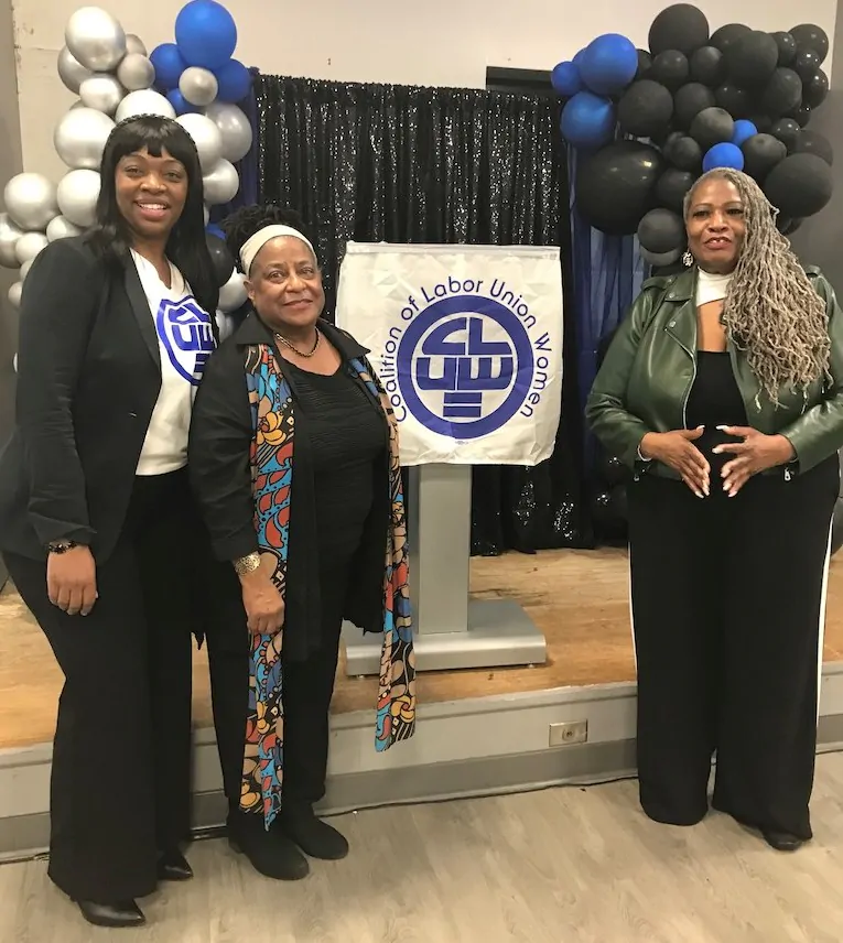 Jovida (middle) stands in the middle alongside two community members at a Coalition of Labor Union Women event. Behind them is a podium with a black backdrop that's decorated with silver, blue, and black balloons.