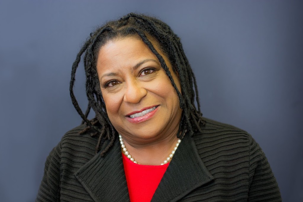Jovida Hill smiles in a close up photograph. She's pictured from the chest up, wearing a black knit blazer over a bright red shirt and a pearl necklace. Her shoulder-length locs frame her face which is set against a navy blue backdrop.